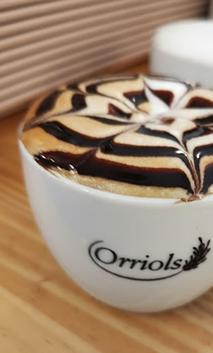 Capuchino en taza serigrafiada con el logo de Orriols, adornado con sirope al estilo arte latte.